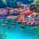Colorful waterfront village with pastel buildings and turquoise water dotted with boats, surrounded by green hillside vegetat